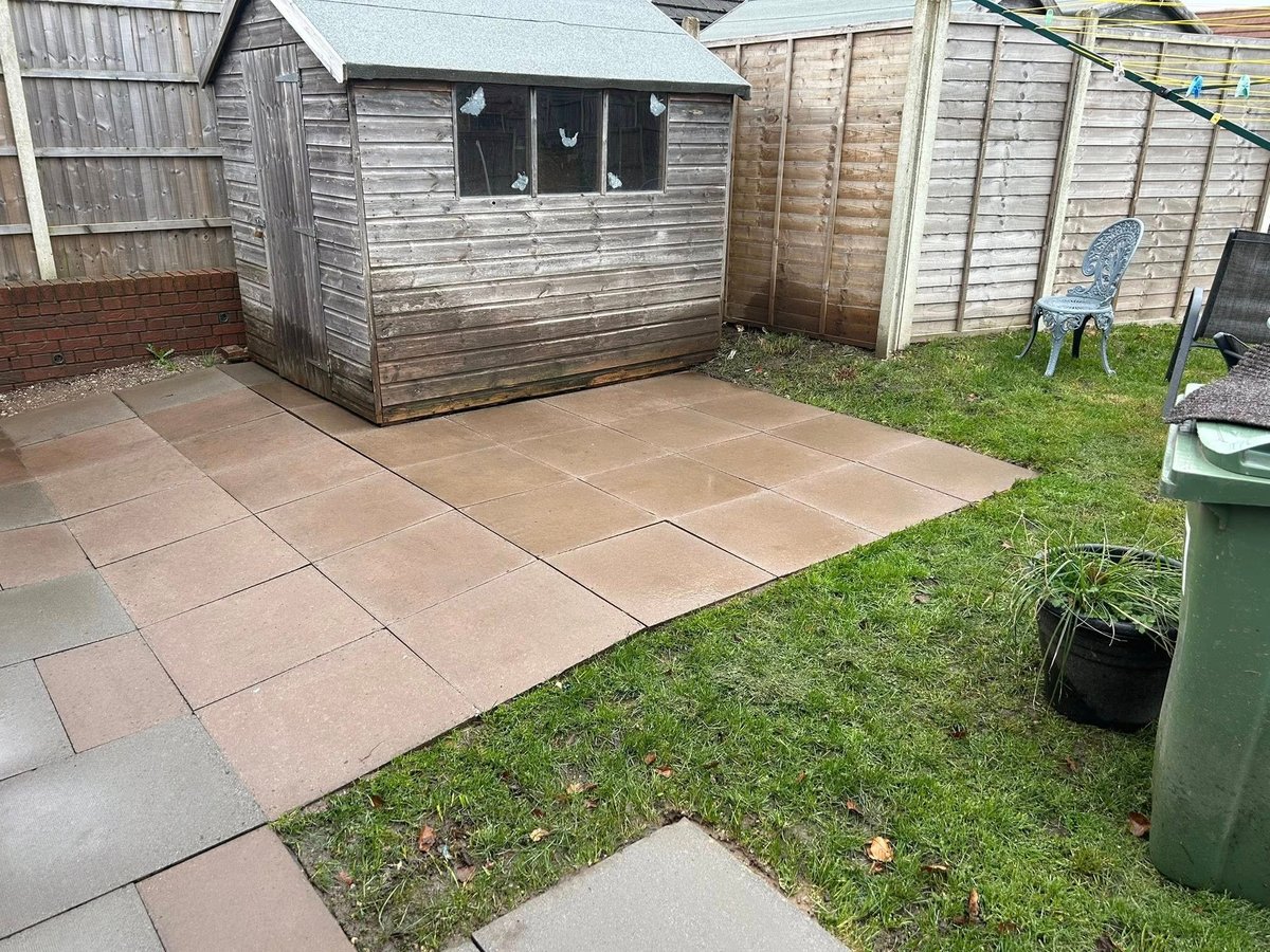 Small wooden garden shed with black window on brown patio surrounded by grass, fences, and wheelie bin in residential garden