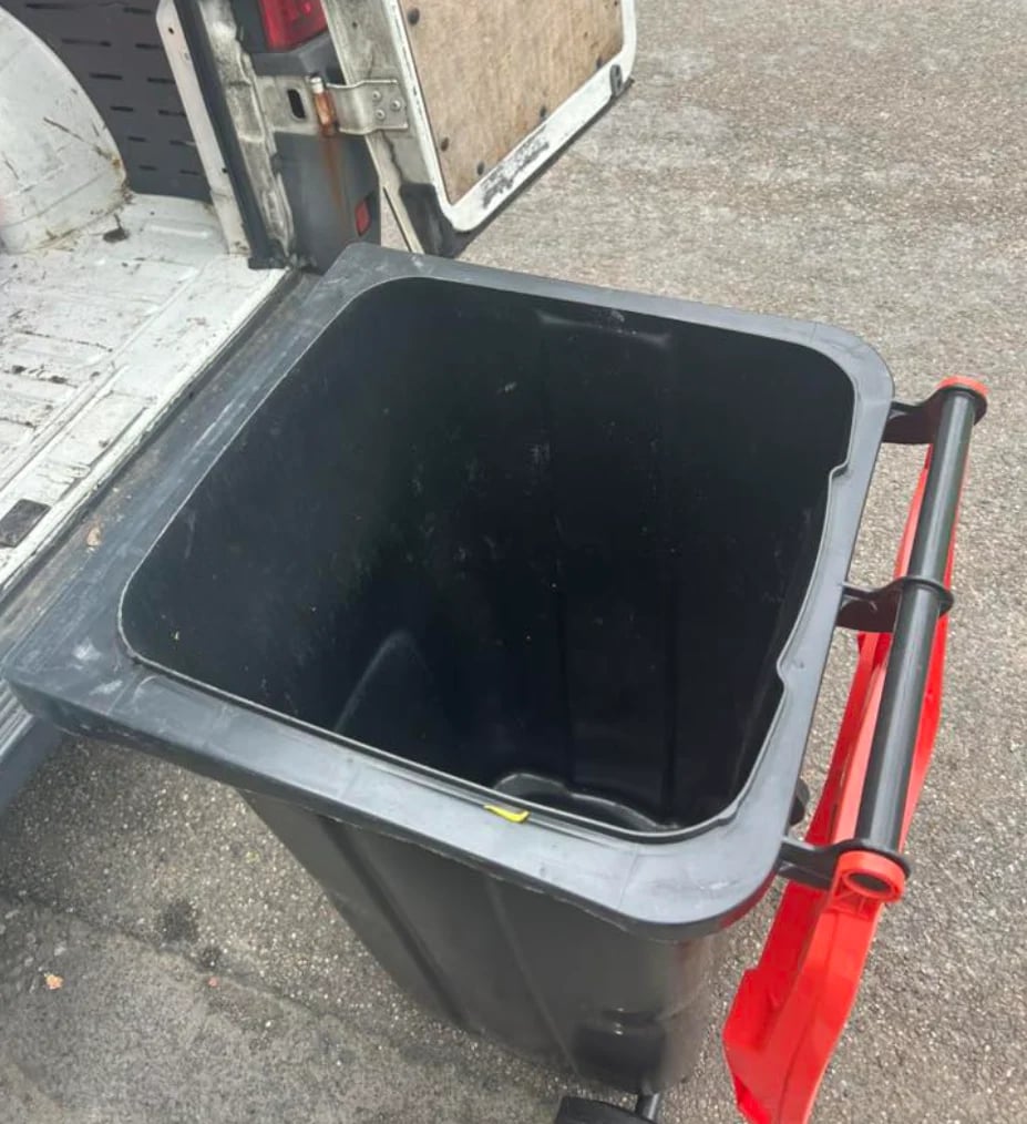 Open black trash bin with red handles on pavement, with a building visible in the background