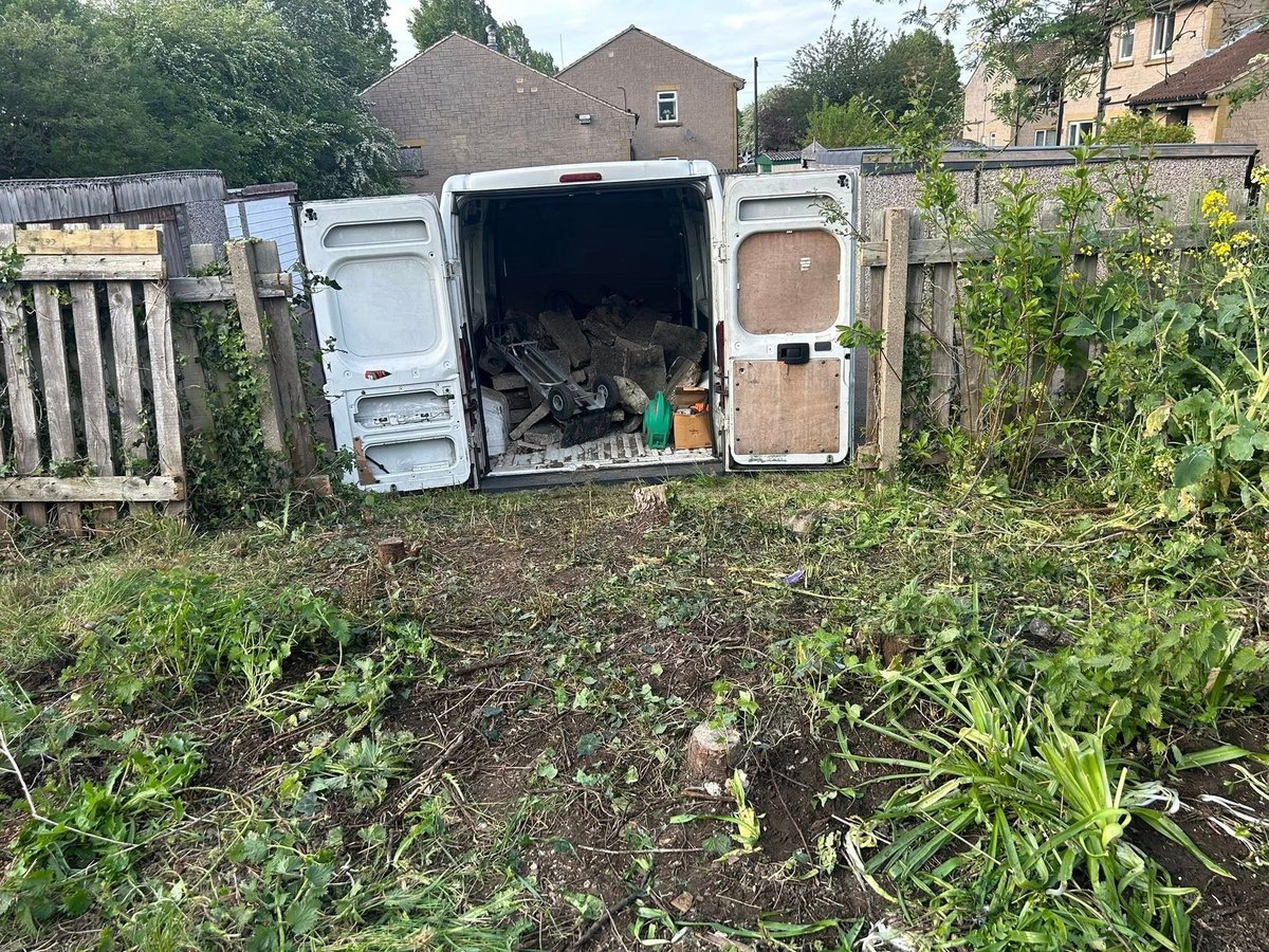 White van with open rear doors in a garden, surrounded by plants and wooden fence