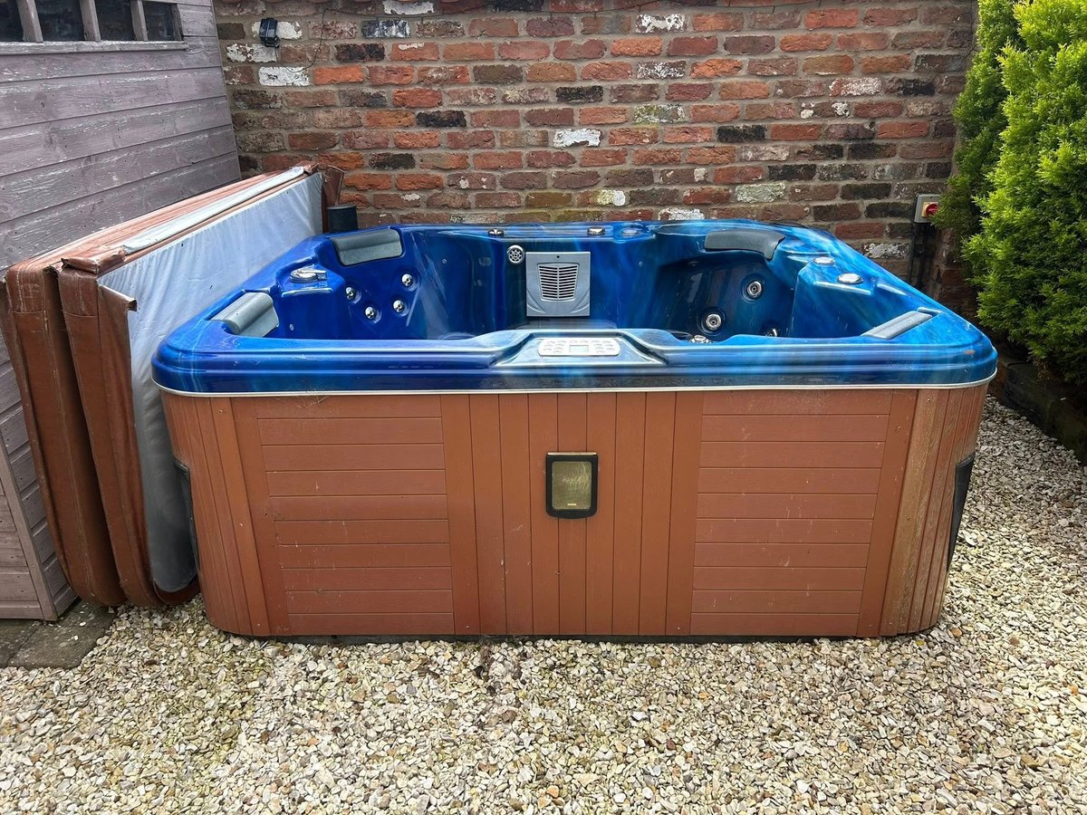 Blue and brown hot tub with cover leaning against brick wall on gravel surface next to green hedge